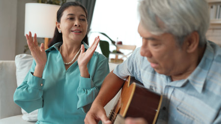 Happy senior man sitting at sofa and playing guitar or music instrument while his wife singing and moving to music. Elder couple spending time together and enjoy doing activity together.の写真素材