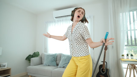 Smiling housekeeper with headphone listen music while cleaning floor. Happy smart caucasian mother mopping floor while dancing and singing to lively song at living room. Mom doing housework. Pedagogyの写真素材