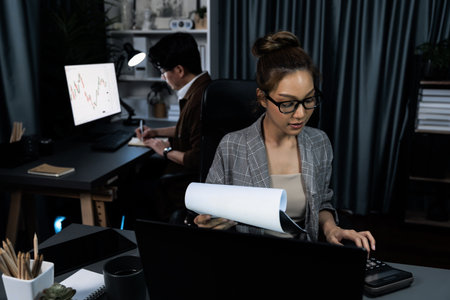 Businesswoman with stressful face looking document paperwork file while coworker trading stock market at back at over late night time at modern office, working on startup company project.の写真素材