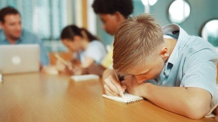 Young smart teenager writing and talking about topic at classroom. Diverse highschool student taking a note in other notebook while sitting with multicultural friends. Creative education. Edification.のeditorial素材