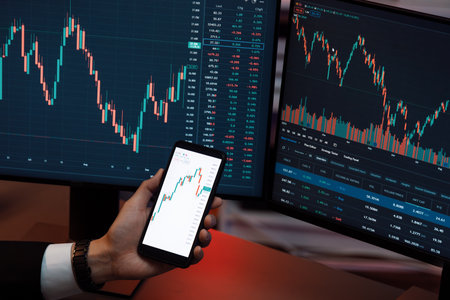 Cropped hand businessman holding smartphone showing dynamic stock market, comparing with financial...