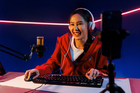 Host channel of smiling beautiful Asian girl streamer playing online game wearing headphone talking with viewers media online recording phone. Esport skilled team players in neon blue room. Stratagem.の写真素材
