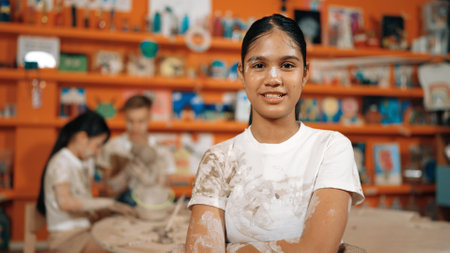 Smiling student looking at camera and crossing arm with confident at workshop while diverse student having pottery class together. Happy girl smile while pose with arm folded.の写真素材