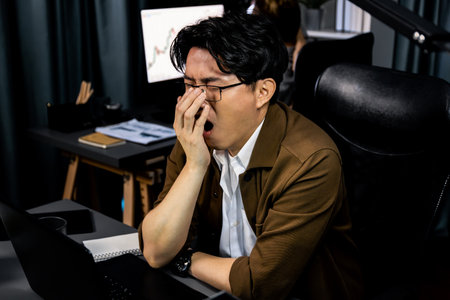 Working businessman feeling extremely sleepy with yawning while working on desk with unfinished project at workplace with coworker trading stock market investment at over late night time. Infobahn.の写真素材