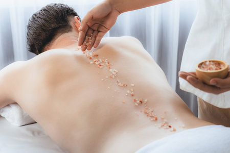 Blissful man customer having exfoliation treatment in luxury spa salon with warmth candle light ambient. Salt scrub beauty treatment in health spa body scrub. Quiescentの写真素材