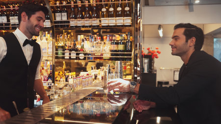 Smart bartender serving drink gin fizz with lemon sliced to VIP guest while waiting friend at luxurious counter bar. Barman providing excellent service at vibrant nightlife party atmosphere.の写真素材