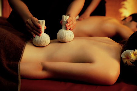 Hot herbal ball spa massage body treatment, masseur gently compresses herb bag on couple customer body. Serenity of aromatherapy recreation in warm lighting of candles at spa salon. Quiescentの写真素材