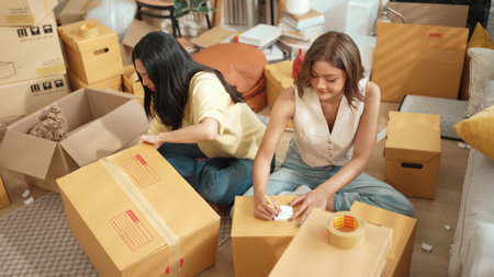 Attractive roommate writing location at parcel while friend packing package at living room. Happy...