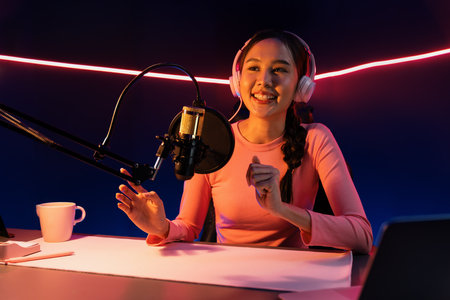 Smiling creative young beautiful Asian influencer talking in channel with positive podcast speech at night time, using laptop for presentation online at pink neon light modern studio.の写真素材