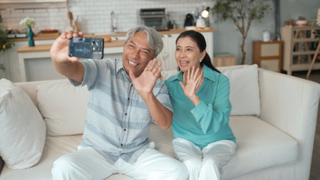 Happy grandfather and grandmother making video call with their son at home. Elder man holding smart phone and sitting at sofa while old woman greeting with cheerful vibe at modern house.の写真素材