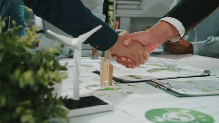This image captures the moment of a business agreement focused on sustainability, featuring a handshake and green energy models, symbolizing collaboration in eco-friendly practices.の写真素材