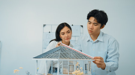 Professional young architect engineer using ruler and pencil measuring model roof length at modern office with blueprint hanged behind. Creative design and teamwork concept.の写真素材