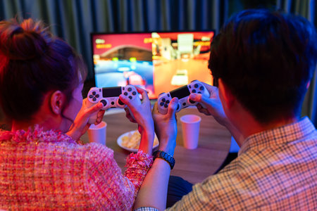 Couple gamer with controlled joystick playing car racing video game together on tv screen with snack at back side view, sitting sofa at neon light colorful living room at modern comfy home. Infobahn.のeditorial素材