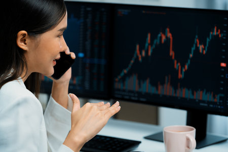 Young beautiful Asian businesswoman calling to broker on phone to analyze stable or higher profit value with profitable each exchange stock market graph on pc screens at modern office. Stratagem.の写真素材