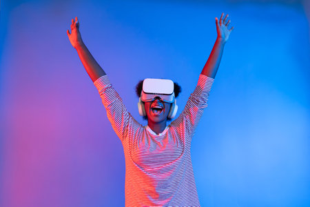 Joyful African woman with VR headset raised hands in excitement on neon studio lighting in virtual reality entertainment with metaverse new world 3D hologram technology.の写真素材