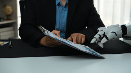 A human and a robotic hand collaborate over documents in a modern workspace, showcasing the integration of technology in business and innovation.の写真素材