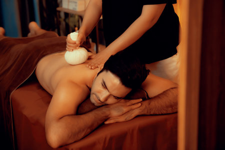 Hot herbal ball spa massage body treatment, masseur gently compresses herb bag on man body. Tranquil and serenity of aromatherapy recreation in warm lighting of candles at spa salon. Quiescentの写真素材