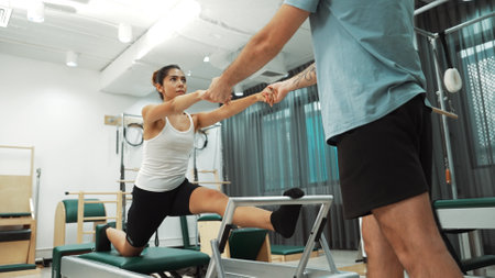 Woman receiving assistance in split stretch exercise during pilates class on trapeze table for flexibility and core training. Student doing split stretch on reformer for improving balance. Habituate.の写真素材