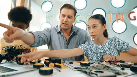 Caucasian student asking teacher about code and pointing at laptop screen. Professional instructor answer girl question while boy repair car model at table with electronic tool placed. Edification.のeditorial素材