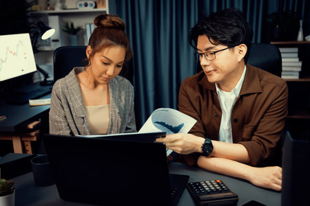 Two managers with stressful face discussing document paperwork file together comparing with report on laptop at over late night time at modern office, working on startup company project job.の写真素材