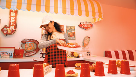 Professional hispanic restaurant worker dancing and standing behind food bar. Attractive waitress with red apron moving to freestyle music while looking at camera. Crazy and funny dance. Regalement.のeditorial素材