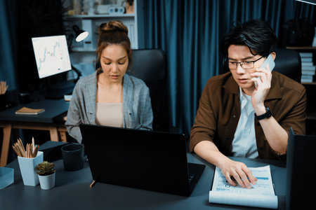 Serious face working on laptop with secretary writing memo on paperwork at over late night time at modern office. Concept of overworked problem project.の写真素材