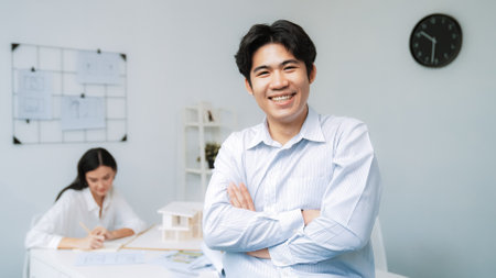 Professional asian businessman crosses arms with confident while young beautiful caucasian colleague drawing a blueprint on meeting table with house model and blueprint at modern office. Immaculate.の写真素材