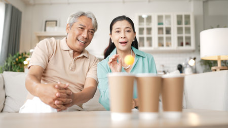 Happy elderly couple enjoying a ball toss game together at home, practicing coordination and cognitive skills for healthy aging. Grandfather and grandmother throwing a ball in the cup.の写真素材