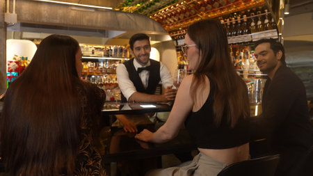 Smart bartender with VIP customer at luxurious bar greeting new beautiful guest at counter in engaging lively conversation at back view in lighting atmosphere at warm nightclub.の写真素材