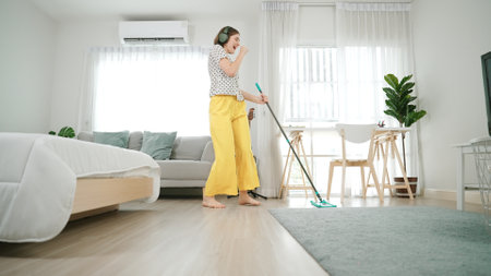 Smiling housekeeper with headphone listen music while cleaning floor. Happy caucasian mother mopping...