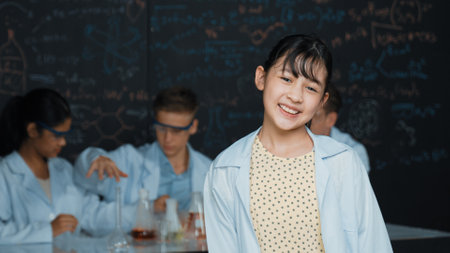 Cute asian scientist smile to camera while diverse group doing experiment. Young happy schoolgirl standing at blackboard with chemical theory written while wearing lab coat at laboratory.の写真素材