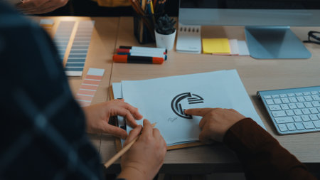 Two creative professionals discuss a logo design, surrounded by color palettes and stationery in a...
