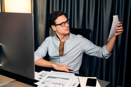 Smart businessman holding paperwork report paragraph dynamic database, looking at pc screen with analyzing global target evaluation value profit company at modern home office at night time.の写真素材