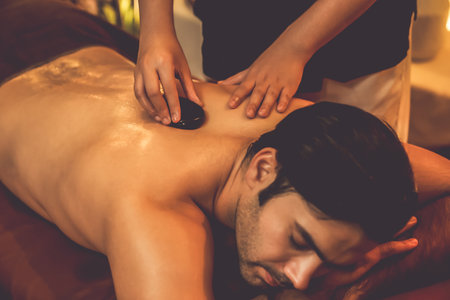 Hot stone massage at spa salon in luxury resort with warm candle light, blissful man customer enjoying spa basalt stone massage glide over body with soothing warmth. Quiescentの写真素材