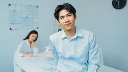 Professional happy businessman smiles during looking at camera. Young beautiful caucasian colleague drawing a blueprint on meeting table with house model and blueprint at modern office.の写真素材
