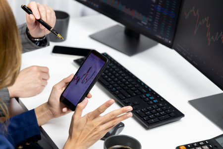 Cropped pointing hand with pen in businessman, checking with holding smartphone by woman investor online website program application with dynamic stock market at home office at back side.の写真素材