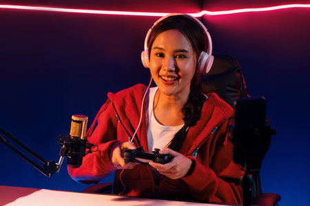 Host channel of smiling Asian girl streamer with joystick playing online game wearing headphones...