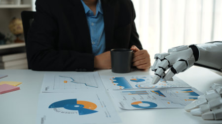 A business professional sits at a modern desk, analyzing charts and graphs with a robotic arm, combining technology with human insight in a futuristic office setting.の写真素材