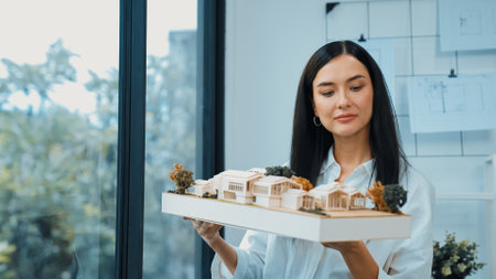 Close up of skilled young beautiful caucasian architect lift house model left side and right sight to check and focus on inspecting mistake point at modern office with blueprint. Immaculate.の写真素材