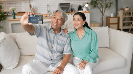Happy grandfather and grandmother making video call with their son at home. Elder man holding smart phone and sitting at sofa while old woman greeting with cheerful vibe at modern house.の写真素材