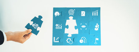 A hand holding a puzzle piece above a completed puzzle featuring various business icons, symbolizing...