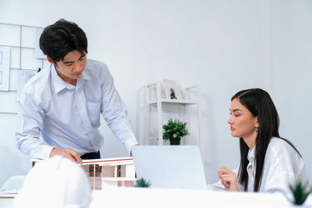 Professional male asian architect using ruler to measure house model length while young beautiful caucasian colleague using laptop to analyzed data on meeting table with house model. Immaculate.の写真素材