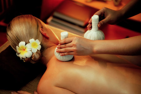 Hot herbal ball spa massage body treatment, masseur gently compresses herb bag on woman body. Tranquil and serenity of aromatherapy recreation in warm lighting of candles at spa salon. Quiescentの写真素材