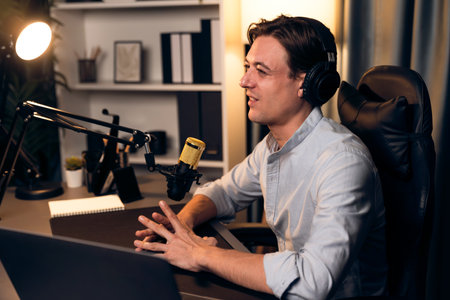 Host channel smart broadcaster talking on live social media application searching on laptop, broadcasting streamer wearing headphone using mic at modern home office studio in warm lighting.の写真素材