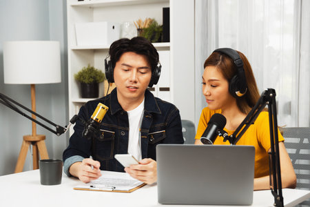 Smiling radio influencer hosts wearing headphones with channel social media online live streaming with talking script note with phone and laptop to listeners at morning at studio record. Infobahn.の写真素材