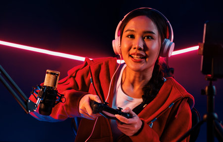 Host channel of smiling Asian girl streamer with joystick playing online game wearing headphones...