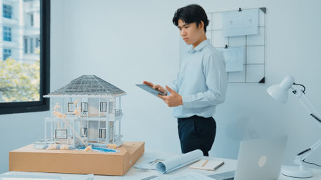 Professional handsome asian project manager using tablet to check house model while inspect house model structure carefully at office with blueprint hanged behind. Creative design. Immaculate.の写真素材