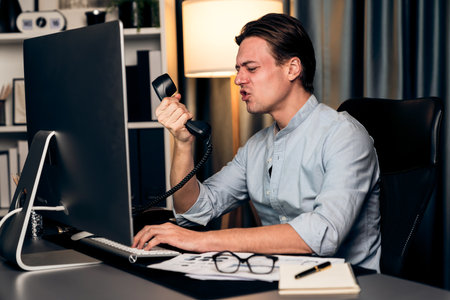 Businessman calling with telephone office argument shouting over talking with partners on dynamic data marketing analysis plan surrounded paper on pc monitor at home office at night.の写真素材