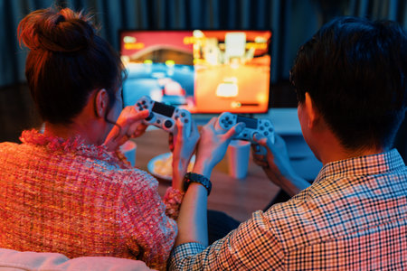 Couple gamer with controlled joystick playing car racing video game together on tv screen with snack at back side view, sitting sofa at neon light colorful living room at modern comfy home.の写真素材
