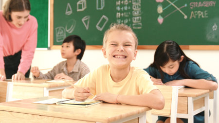Caucasian smart child smiling at camera while doing classwork at classroom while happy multicultural student doing test or writing note in classroom at elementary class. Education concept. Pedagogy.の写真素材